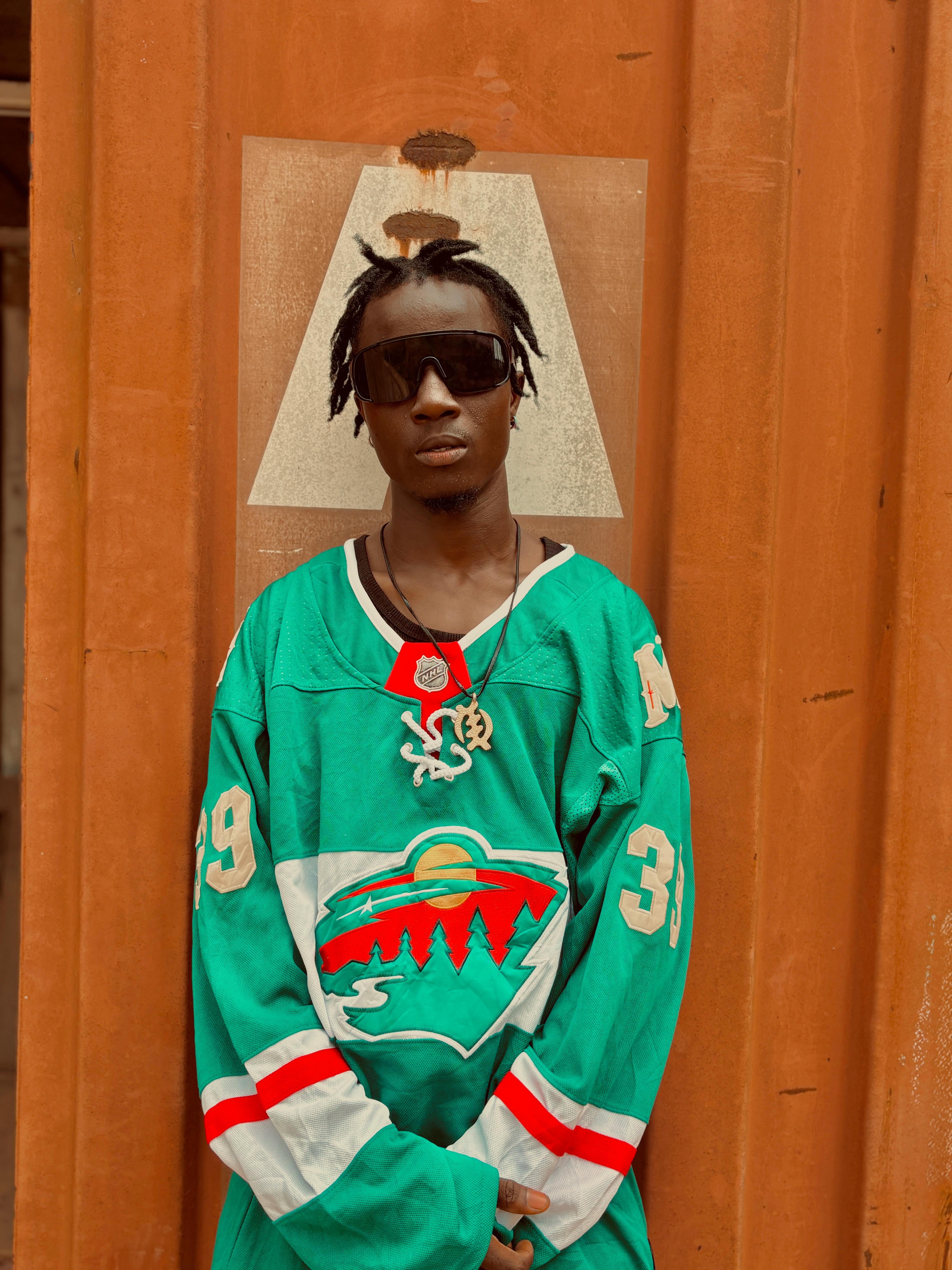 Stylish young man in sunglasses wearing a green hockey jersey against a rusty metal background.