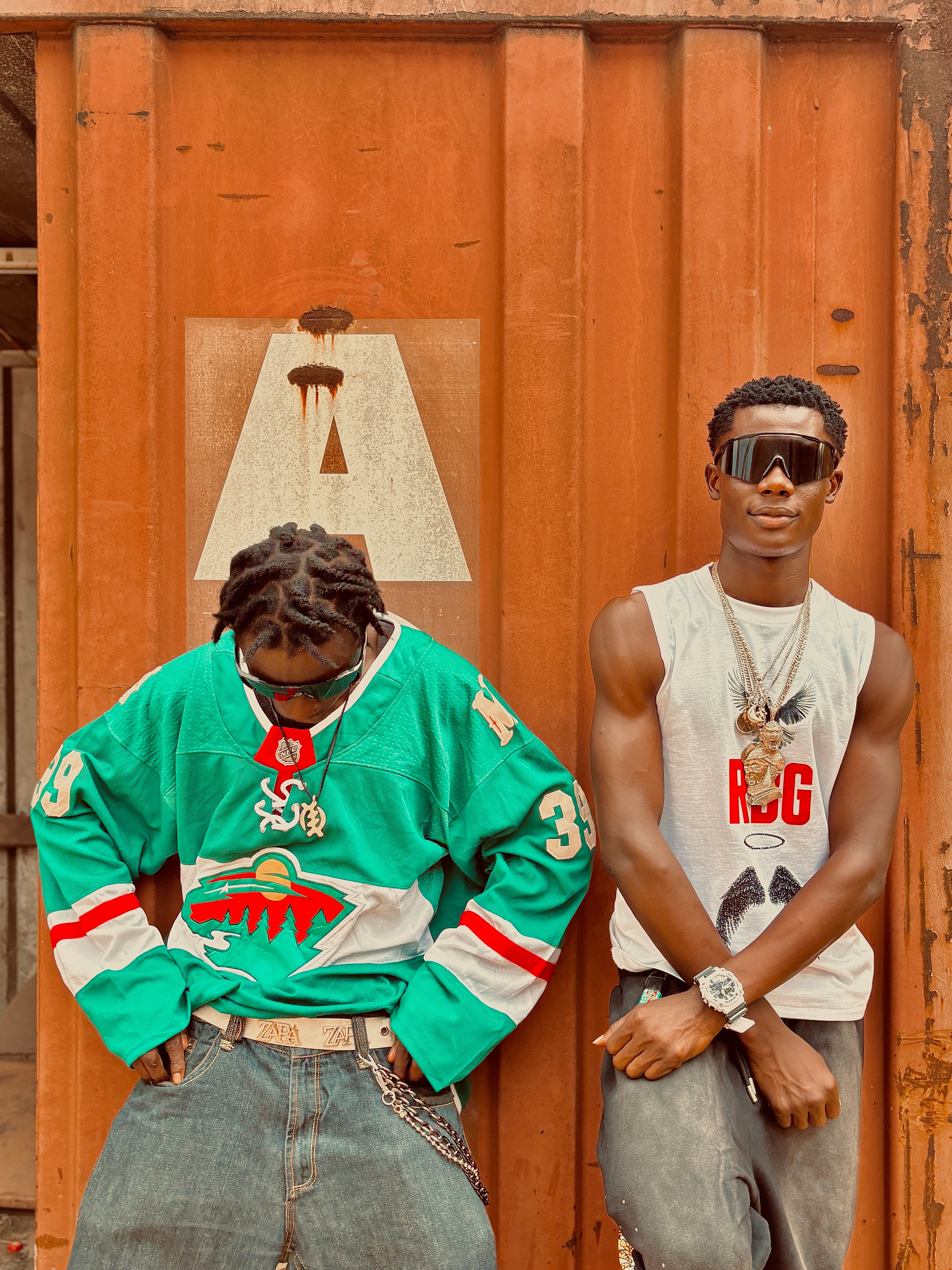 Two stylish young men in trendy streetwear pose confidently against a rustic background.