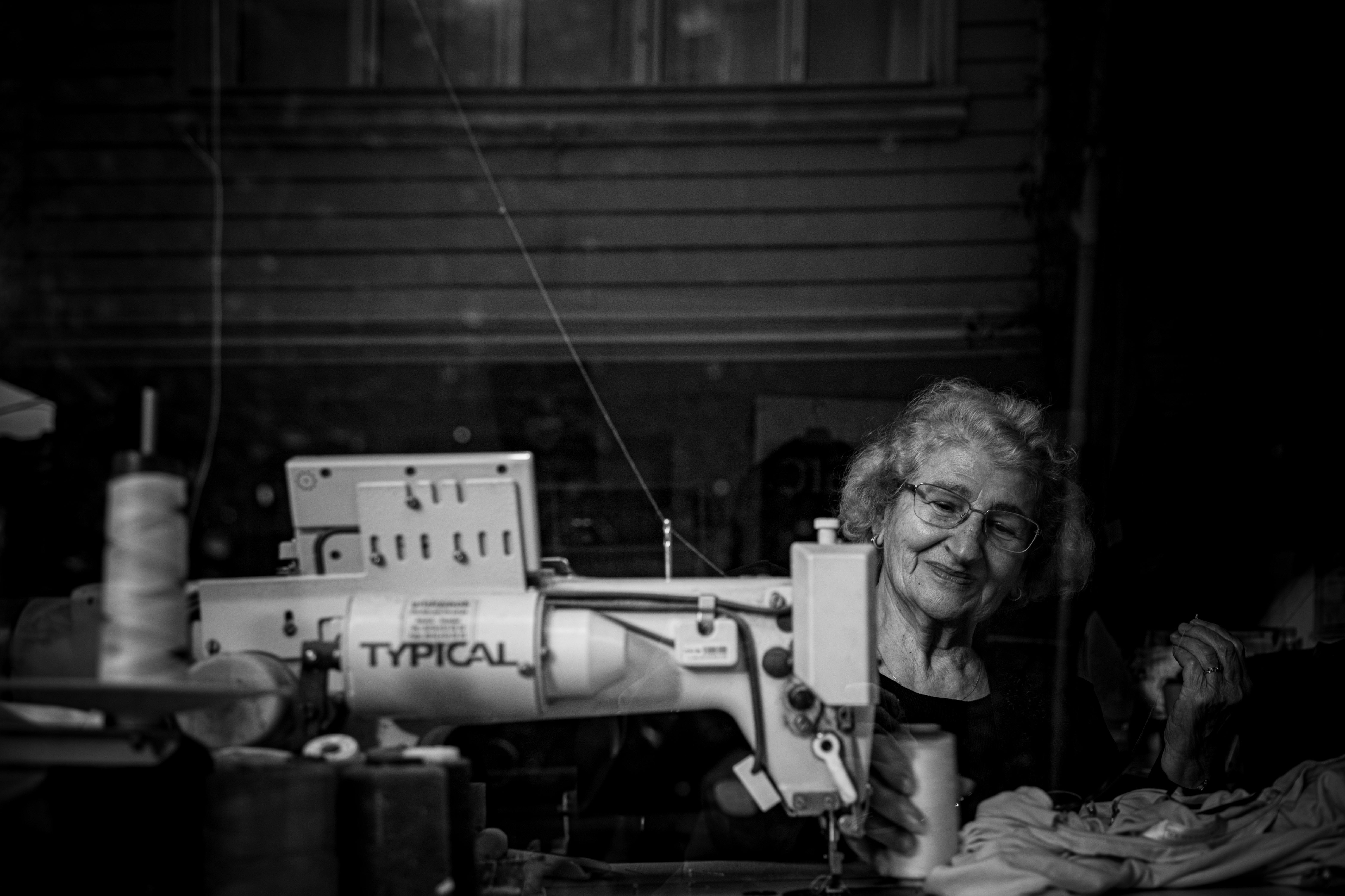 Black and white portrait of an elderly woman sewing, seen through a reflective window in Istanbul.