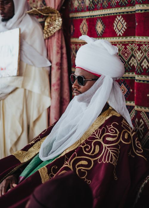 Free African man wearing ornate traditional attire and turban, seated with sunglasses, showcasing cultural elegance. Stock Photo