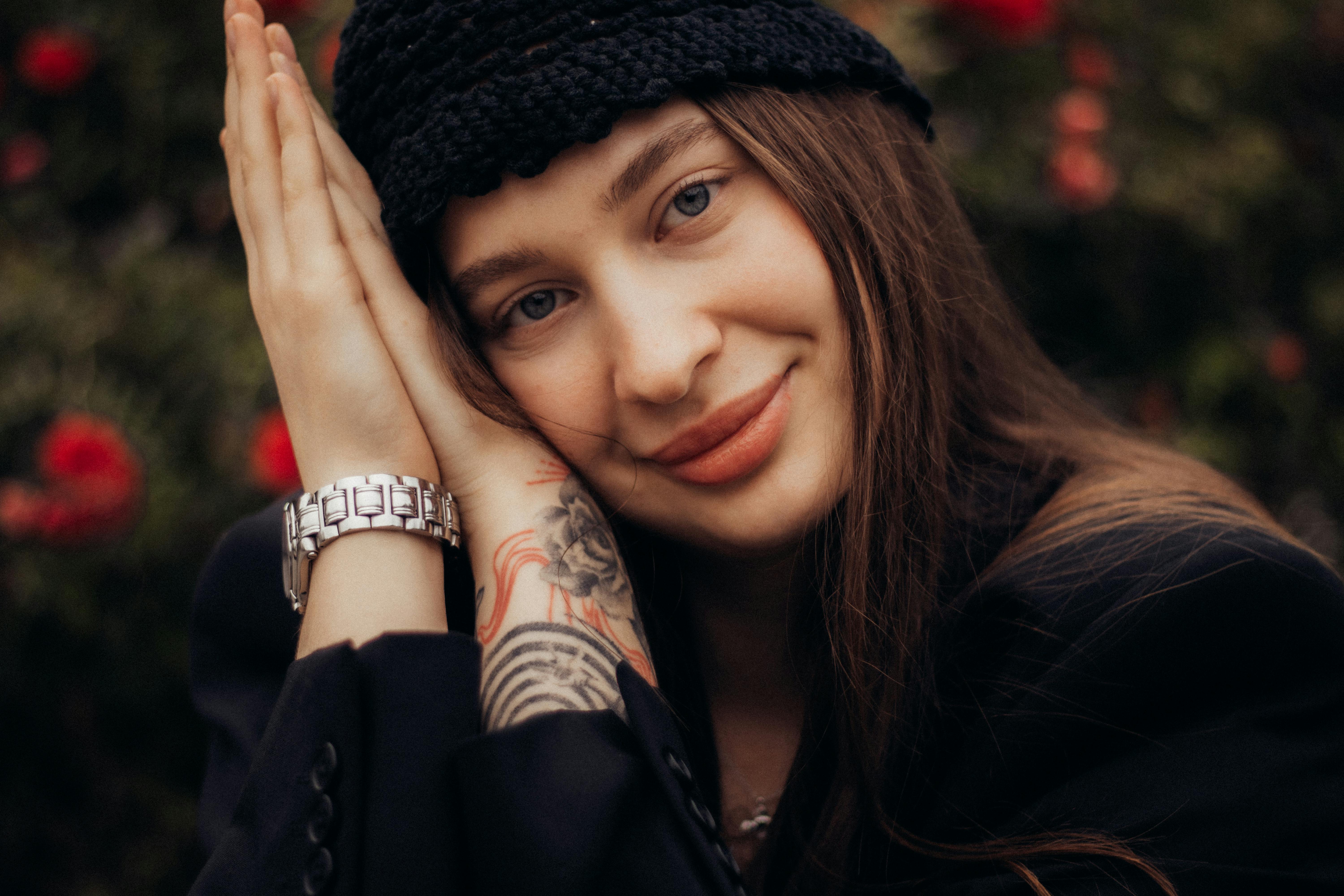 무료 검은 모자와 꽃 문신을 한 젊은 여성이 미소 짓는 클로즈업 초상화가 흐릿한 꽃 배경과 대조를 이루고 있습니다. 스톡 사진