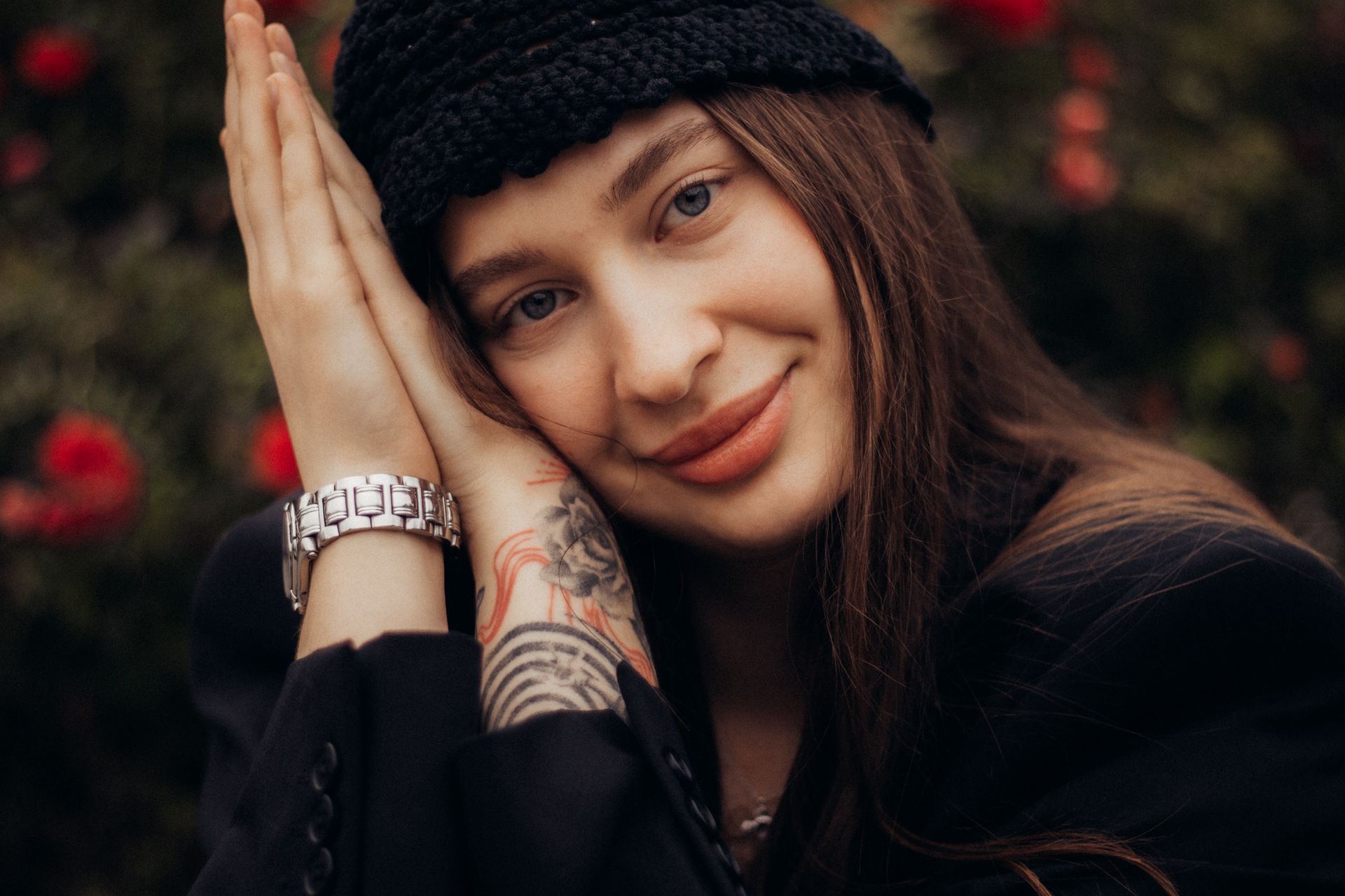 A close-up portrait of a young woman smiling, with a black hat and floral tattoo, set against a blurred floral background.
