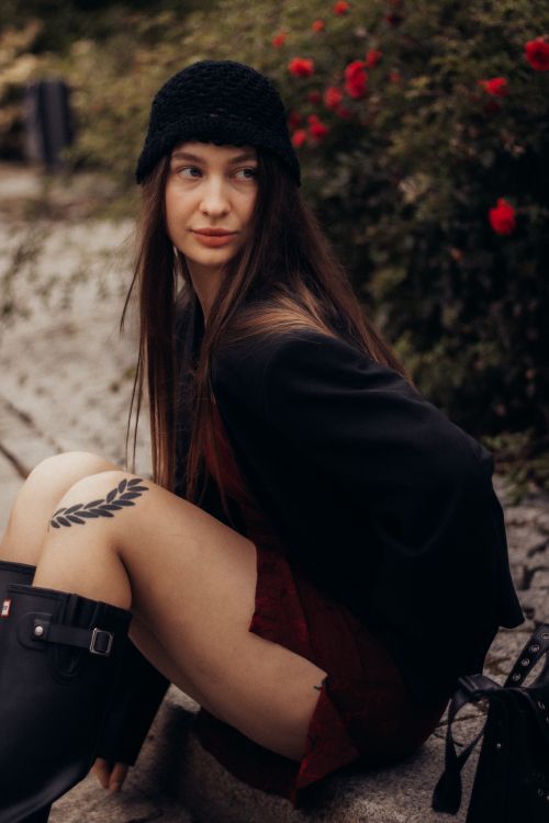 Free A fashionable woman sitting outdoors, wearing a black beanie and boots, surrounded by greenery. Stock Photo