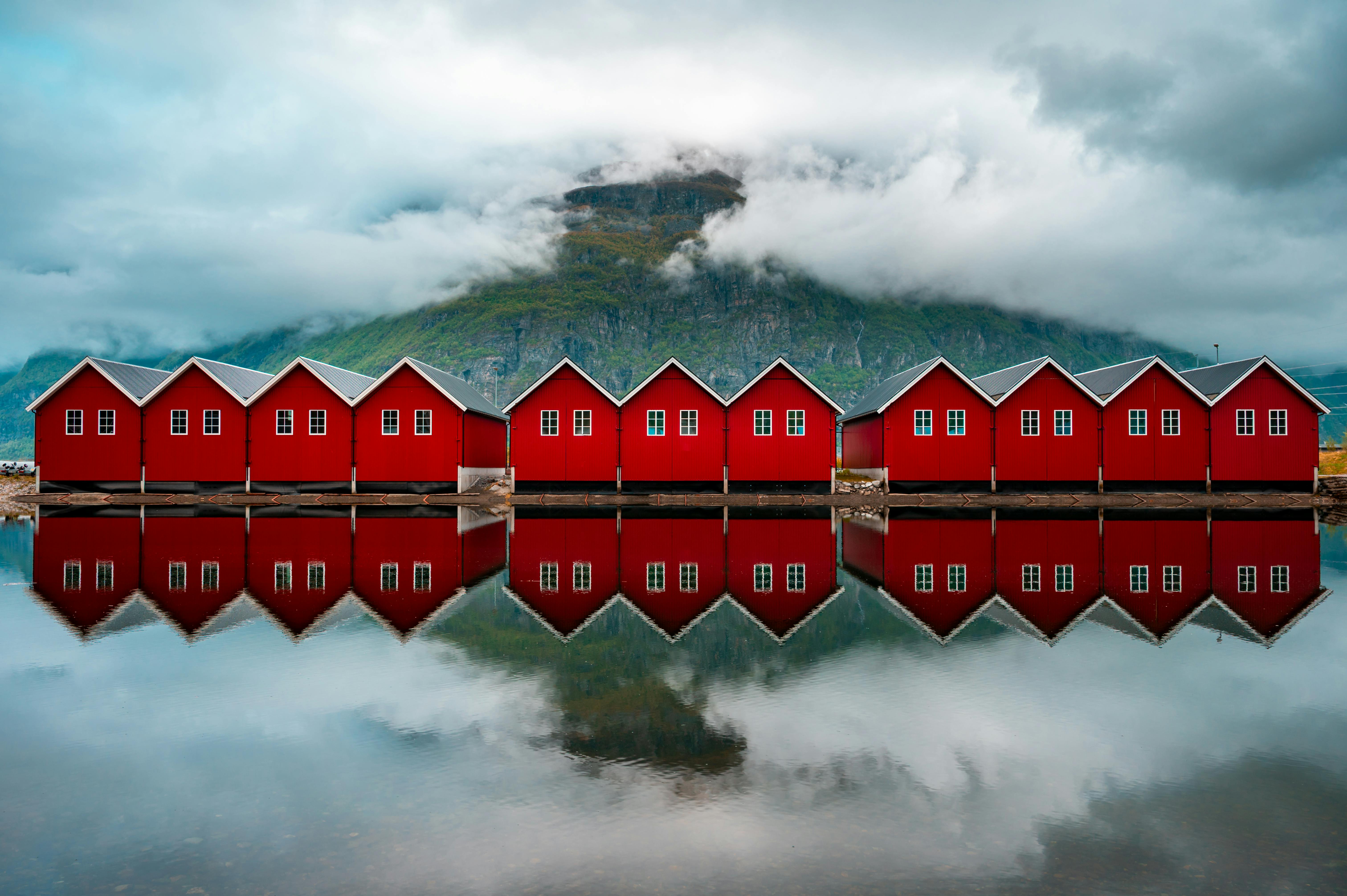 Vibrant red cabins reflect on a serene Norwegian fjord, surrounded by majestic mountains and clouds.