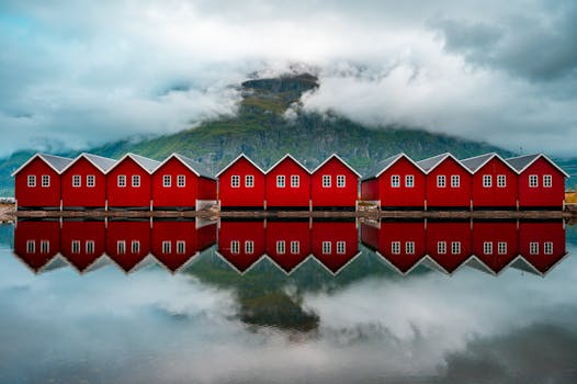 Vibrant red cabins reflect on a serene Norwegian fjord, surrounded by majestic mountains and clouds.