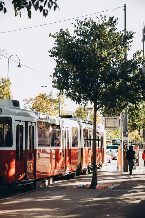 Free A classic red tram on a sunny day in Vienna, Austria, with trees and a walking figure. Stock Photo