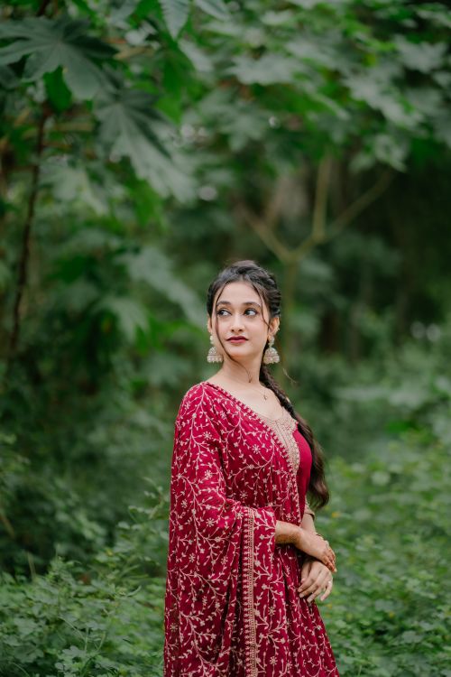 Free A woman in traditional red saree poses against a lush green backdrop. Stock Photo