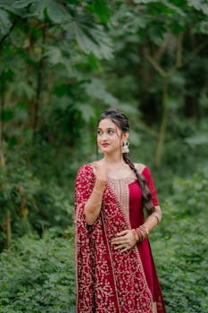Young woman in traditional red attire posing gracefully outdoors in lush greenery.
