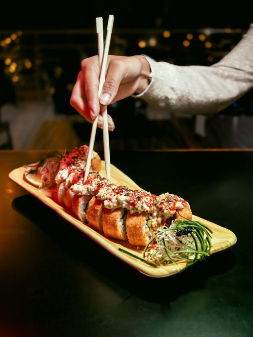 Free A hand using chopsticks to pick up sushi rolls on a wooden platter, captured indoors. Stock Photo