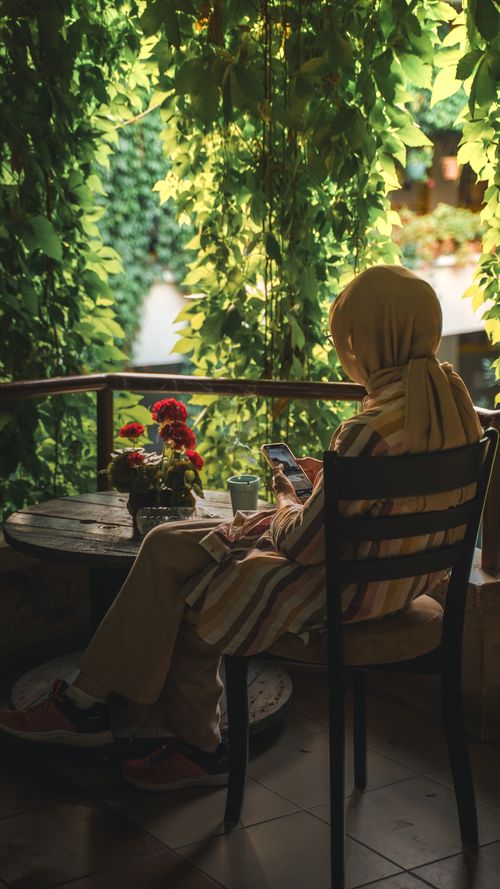 Free A woman in a hijab sits in a leafy cafe in Bursa, Türkiye, using her phone. Stock Photo