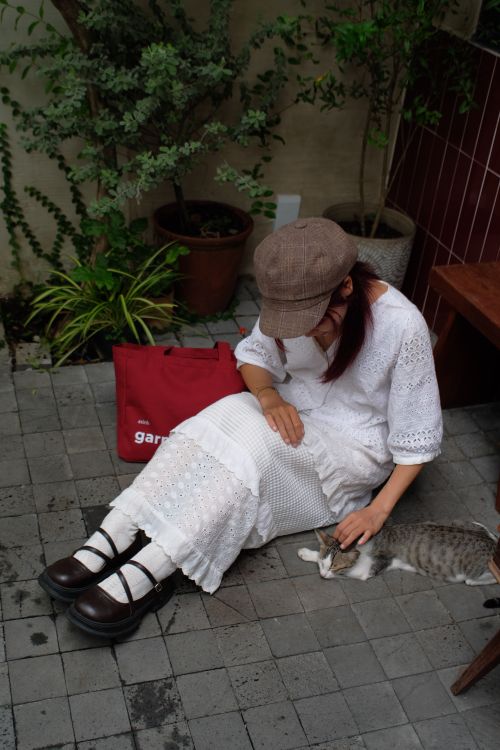 Free Woman in white dress and cap sitting on patio with a cat and plants. Stock Photo