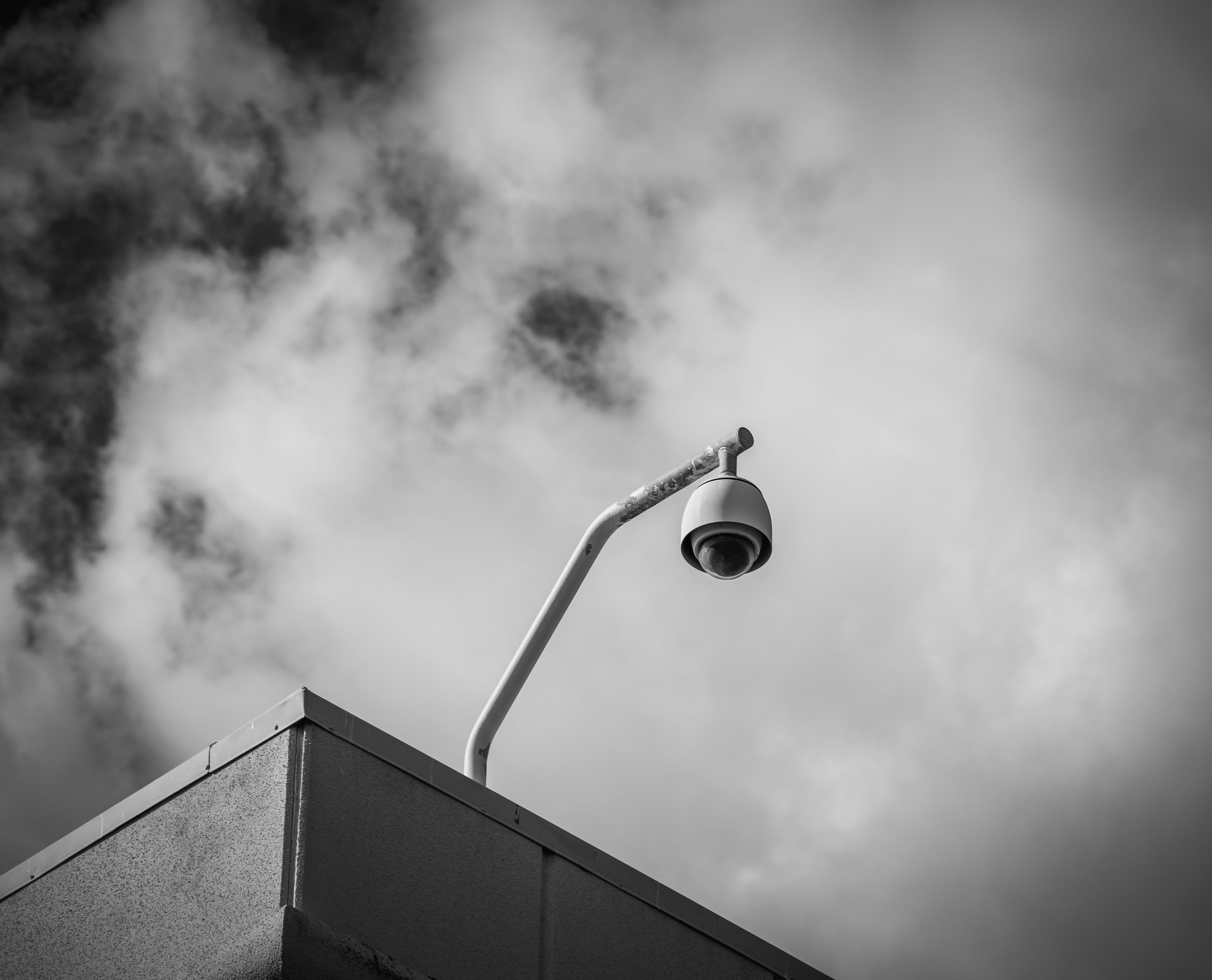 Black and white photograph of a security camera mounted on a building corner against a cloudy sky.