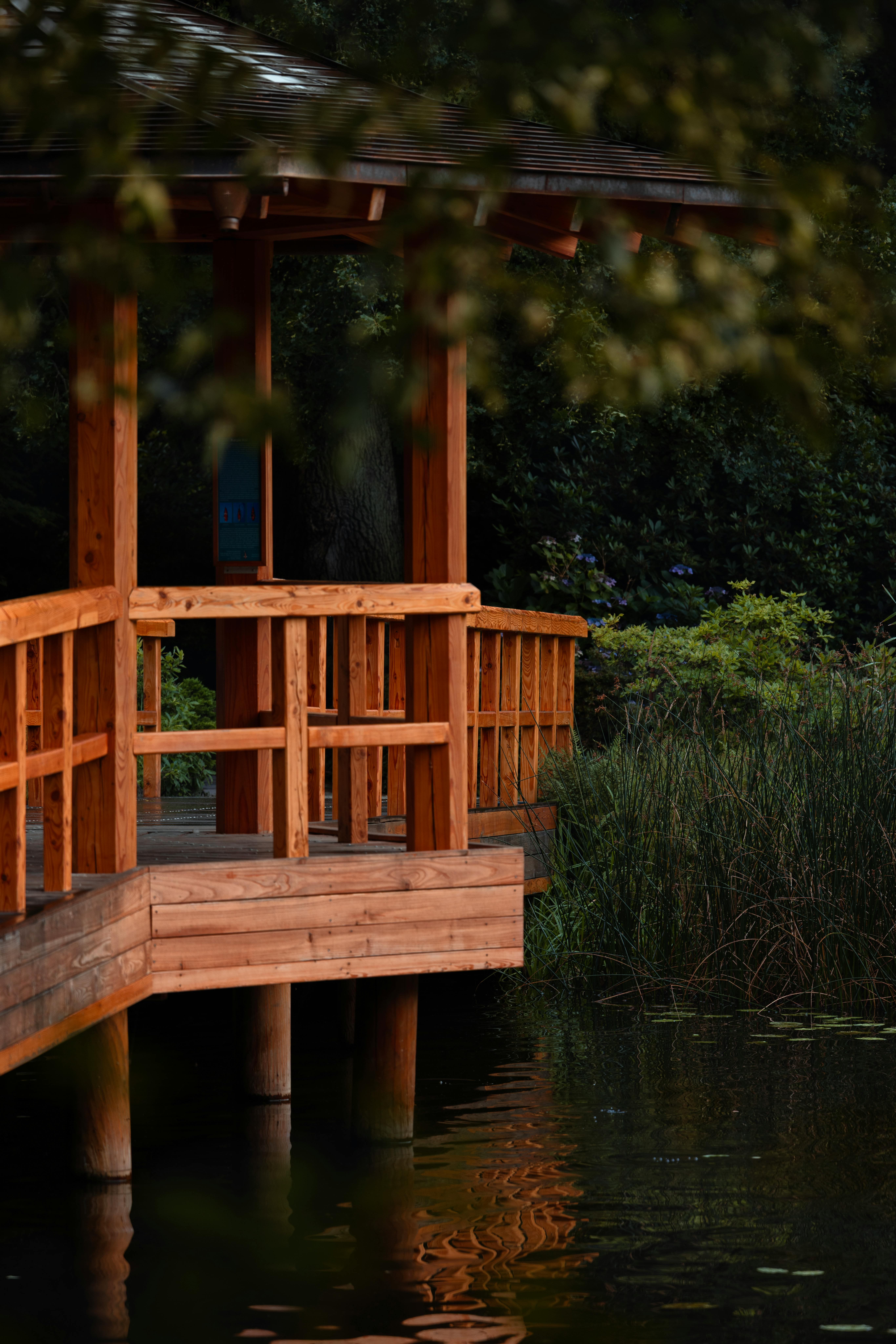 A tranquil wooden gazebo beside a calm pond, surrounded by lush greenery in a peaceful outdoor setting.