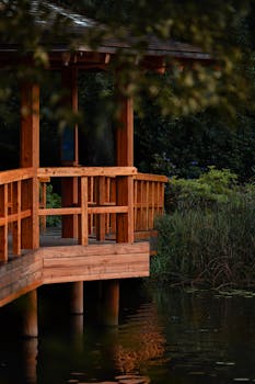 A tranquil wooden gazebo beside a calm pond, surrounded by lush greenery in a peaceful outdoor setting.
