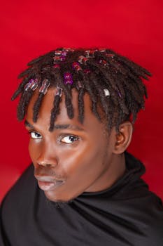Close-up portrait of a stylish young man with unique beaded hairstyle against a vibrant red backdrop.