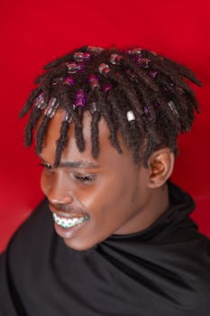 Colorful portrait of a stylish man with beaded dreadlocks and braces, smiling against a vibrant red background.