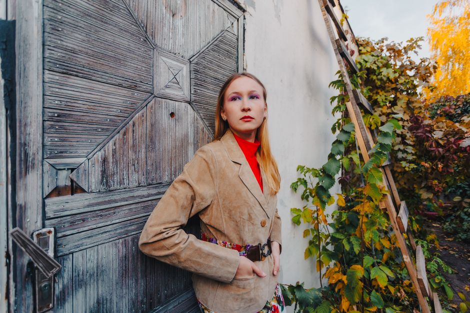 A woman in a tan suede jacket over a red turtleneck stands beside a weathered wooden door surrounded by fall foliage, showing how to mix suede and fine knit textures for fall