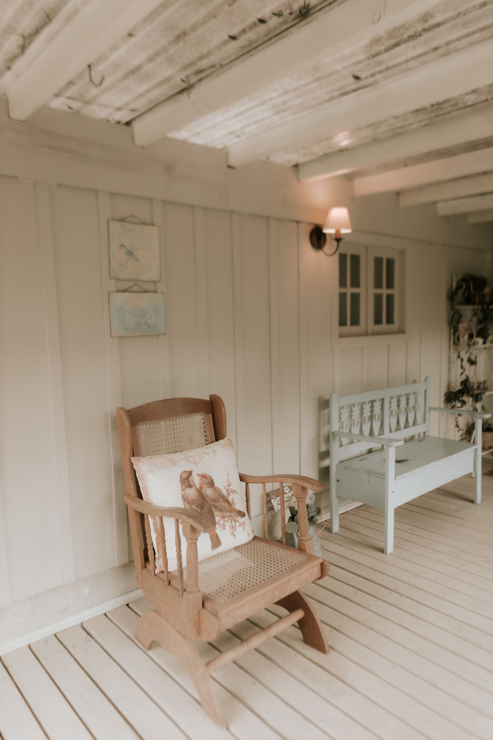 Pale painted porch boards beneath a wooden rocking chair and a light-colored bench.