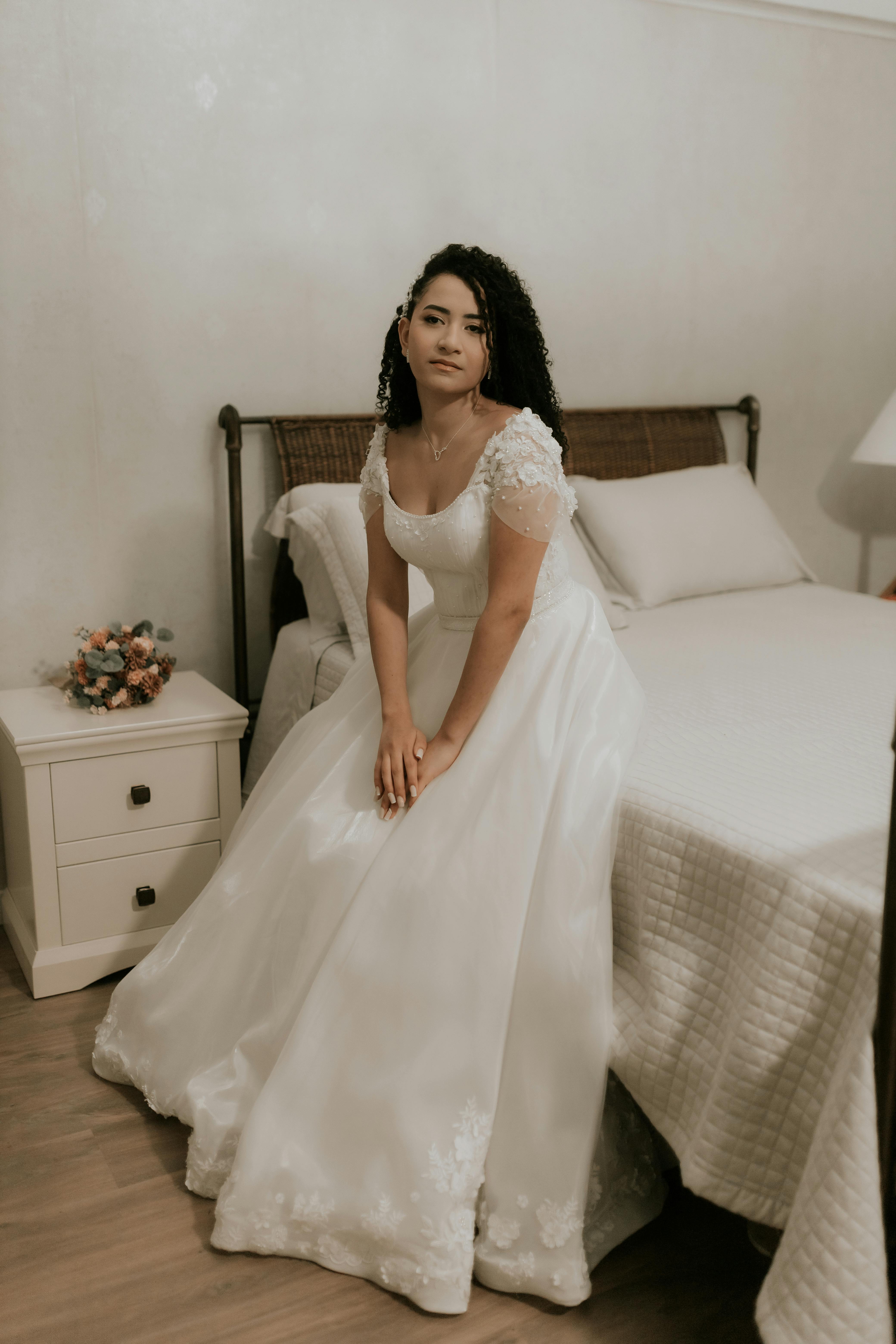 A bride in a classic white wedding dress sits gracefully on a bed, indoors.