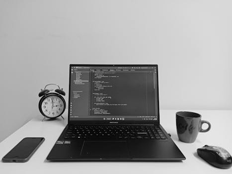 Black and white workspace with a laptop showing code, alarm clock, and coffee mug.