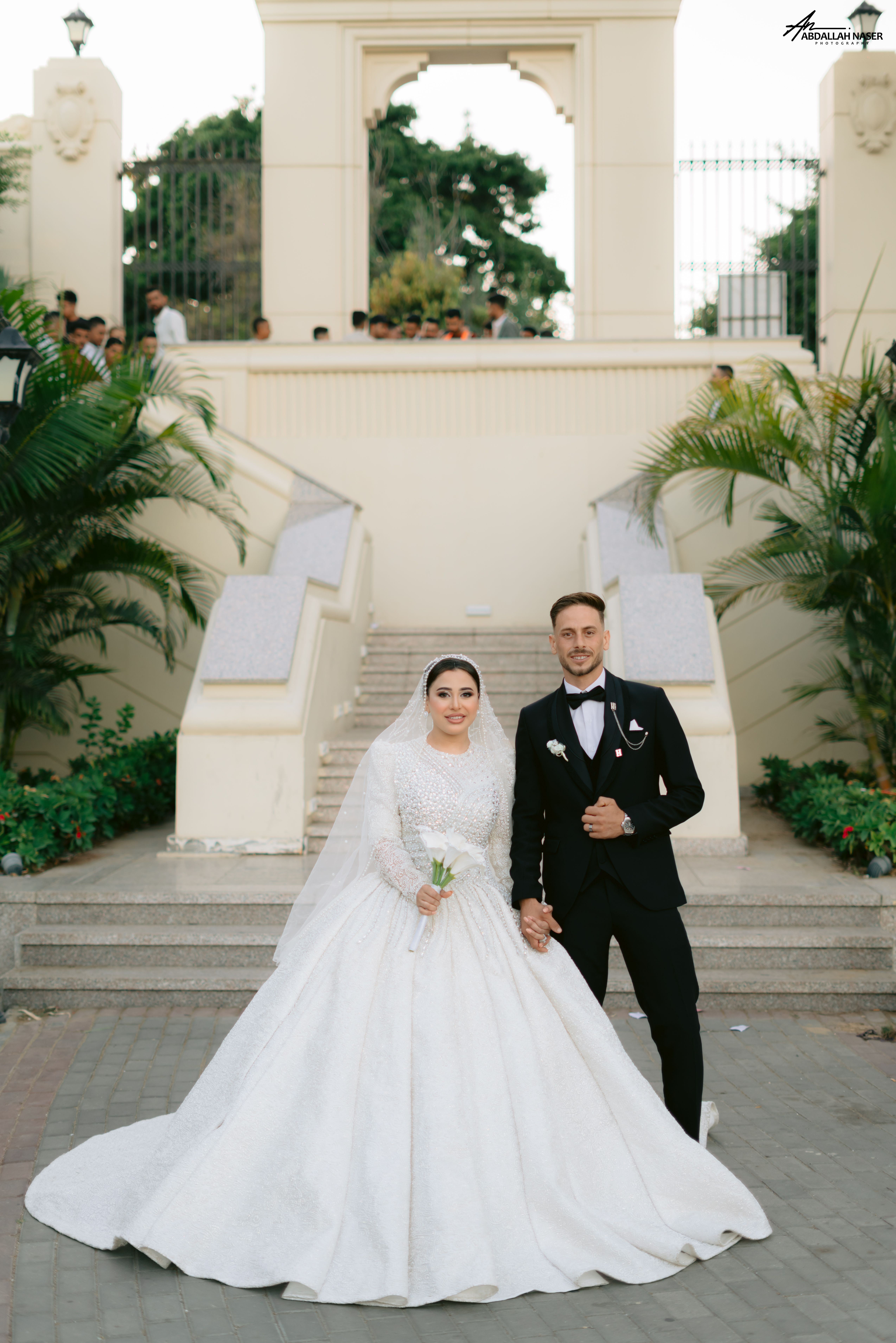 A bride and groom pose joyfully in front of a grand staircase on their wedding day.