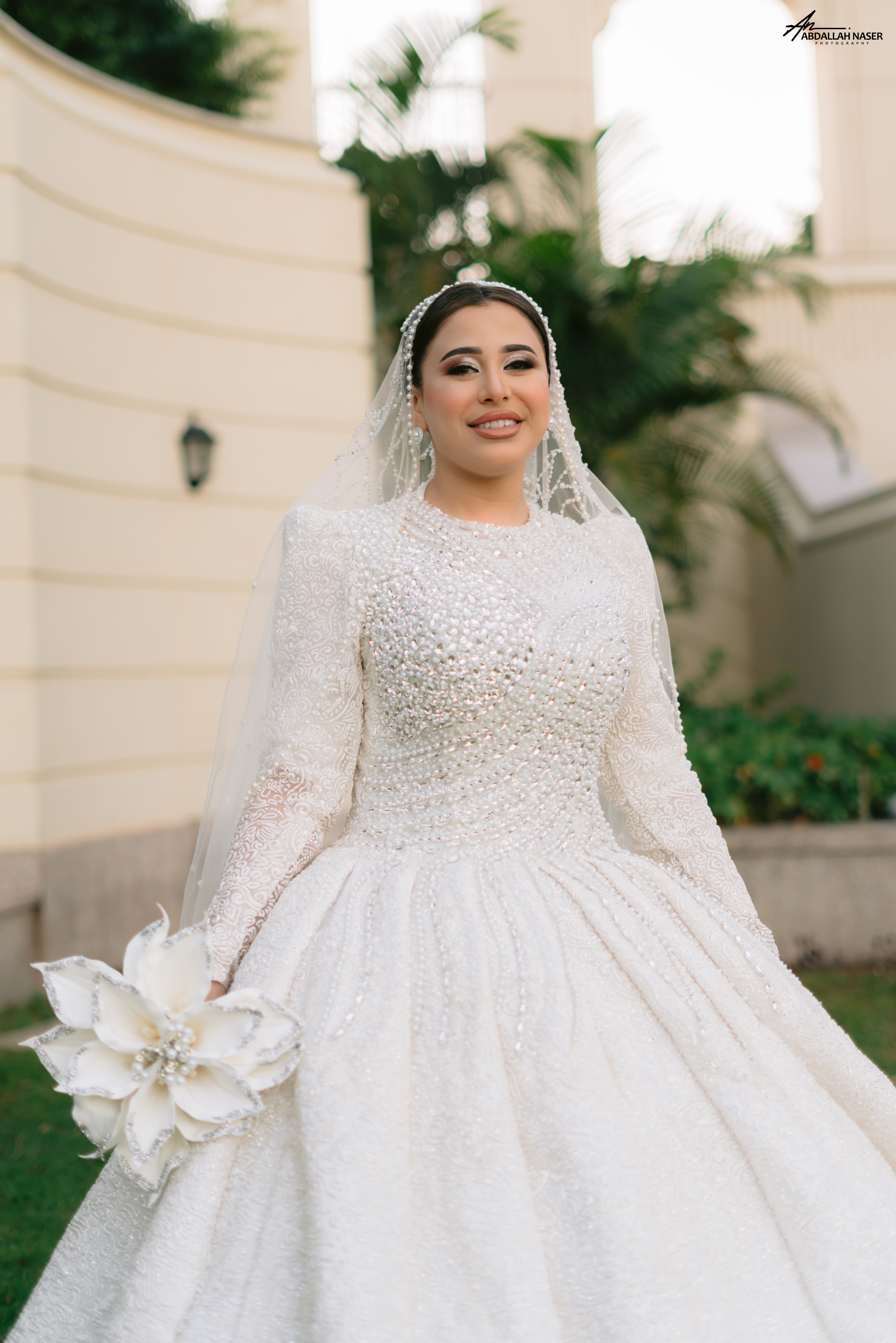 Stunning bride in a detailed white gown holding a bouquet outdoors against a grand architectural backdrop.