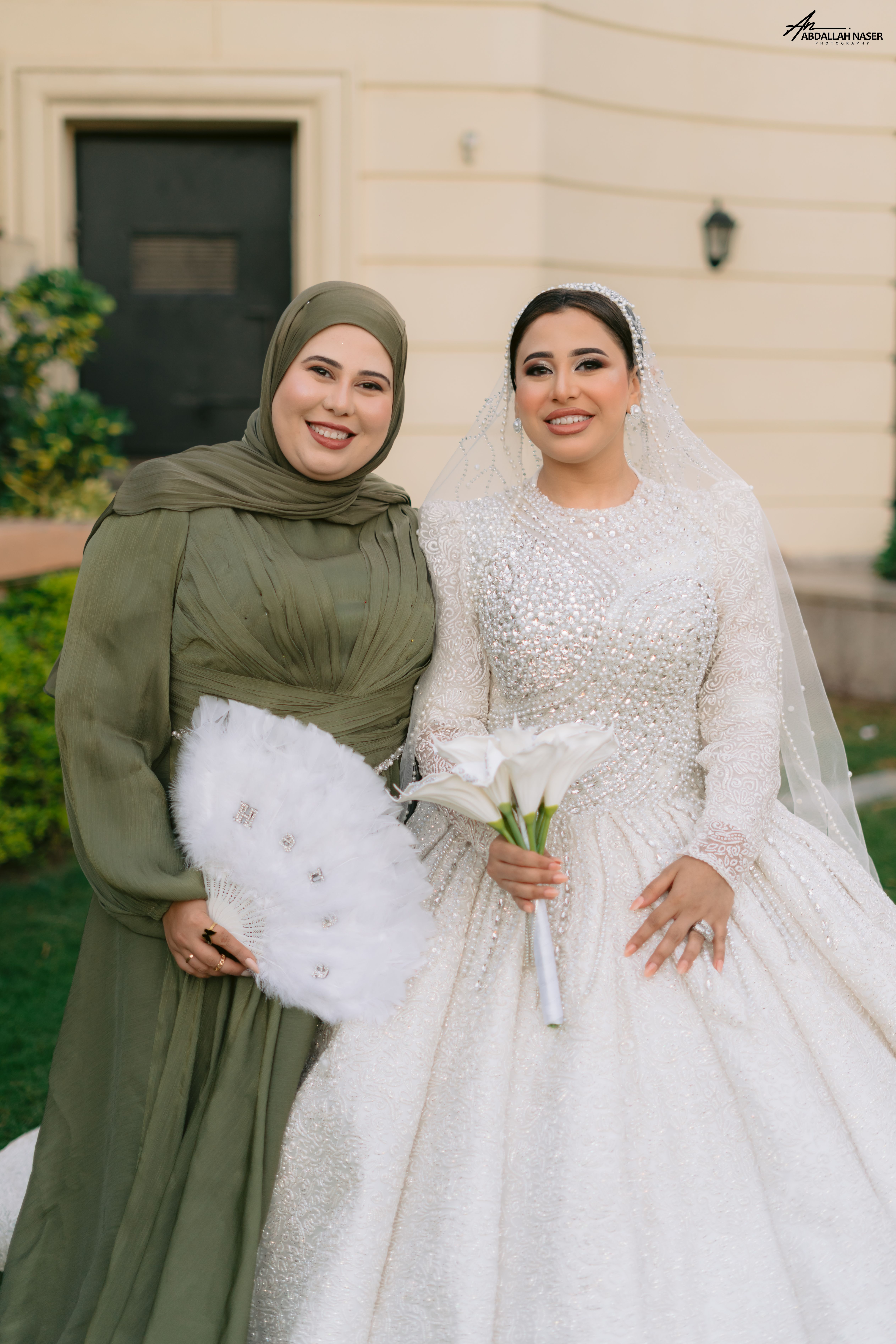 A bride in a luxurious gown poses with a guest outdoors.