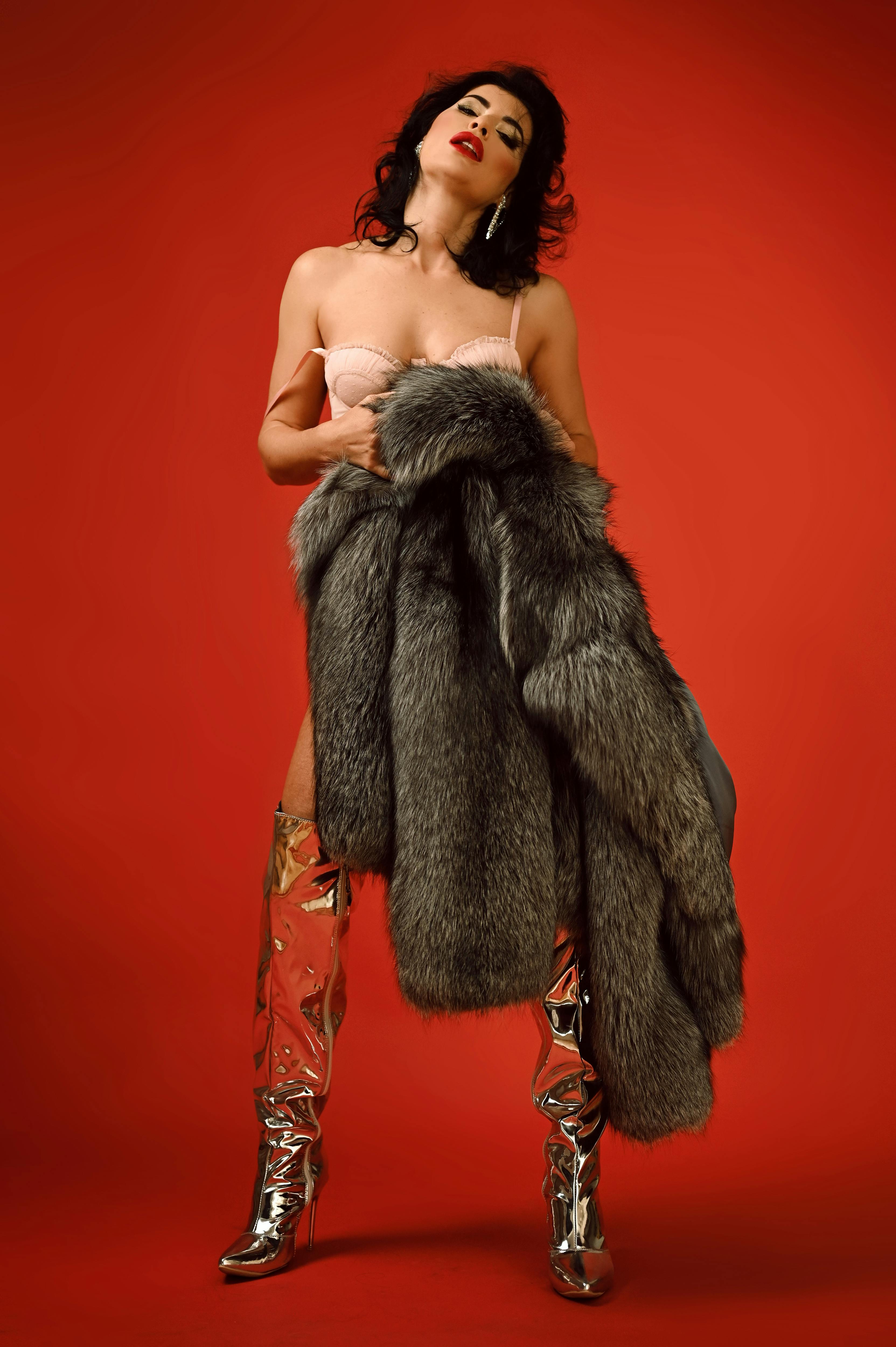 Fashion model poses boldly in fur coat and silver boots against red backdrop.