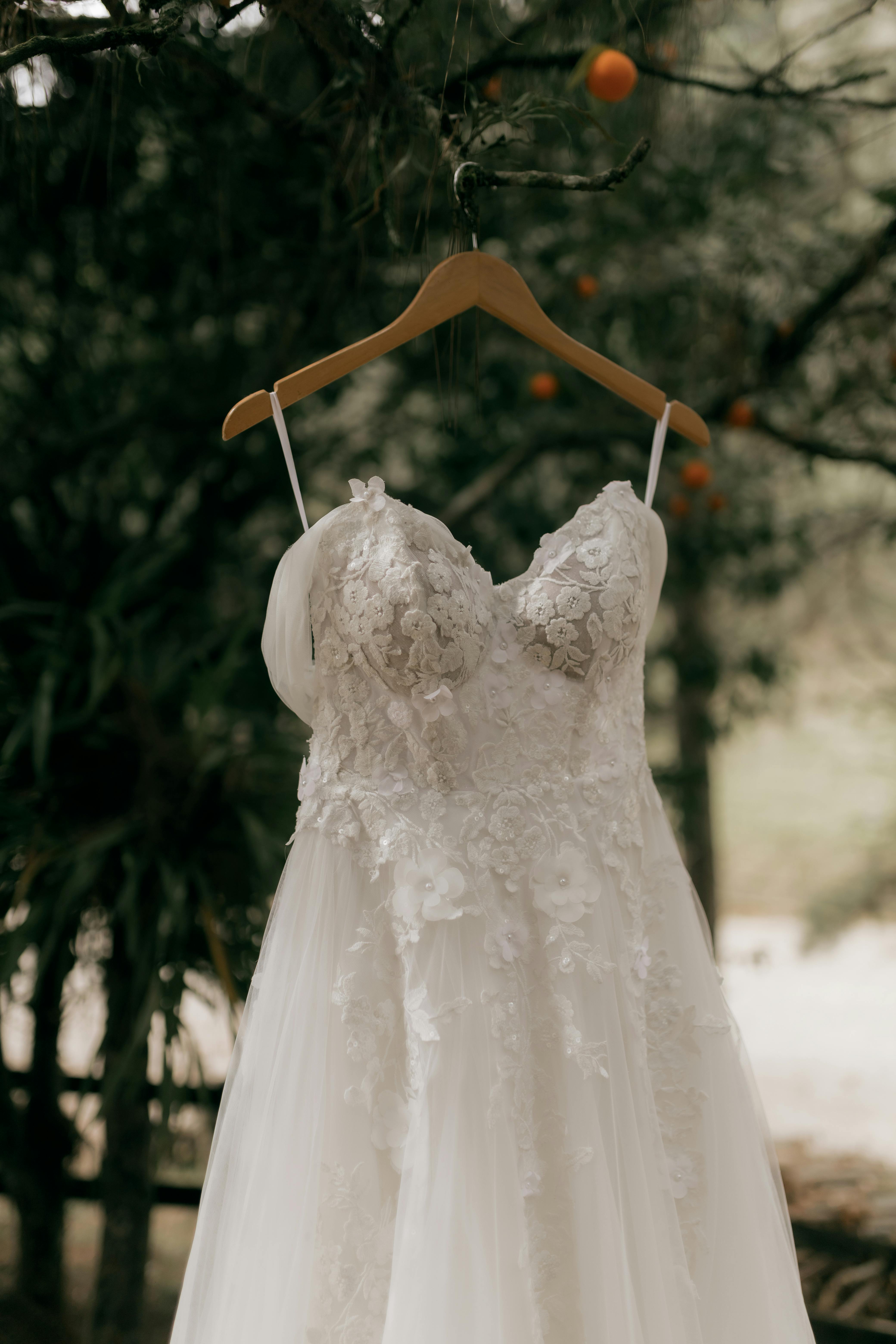 A beautiful lace wedding dress hanging on a wooden hanger in an outdoor setting.
