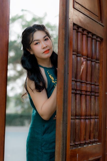 Young woman in a green dress leaning by a carved wooden doorway outdoors.