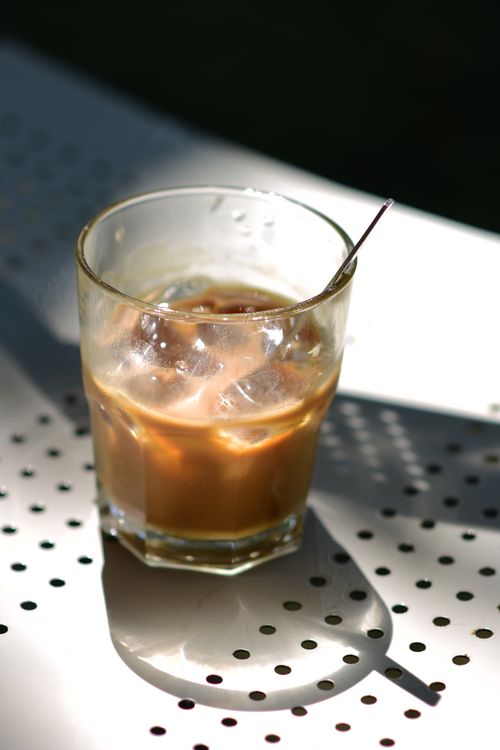 Free A glass of iced coffee with a spoon sitting on a perforated metal table in bright sunlight. Stock Photo
