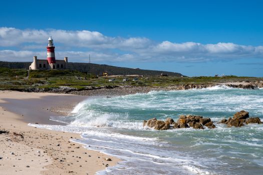 A scenic view of Cape Agulhas Lighthouse with waves crashing on the rocky beach under a bright blue sky.