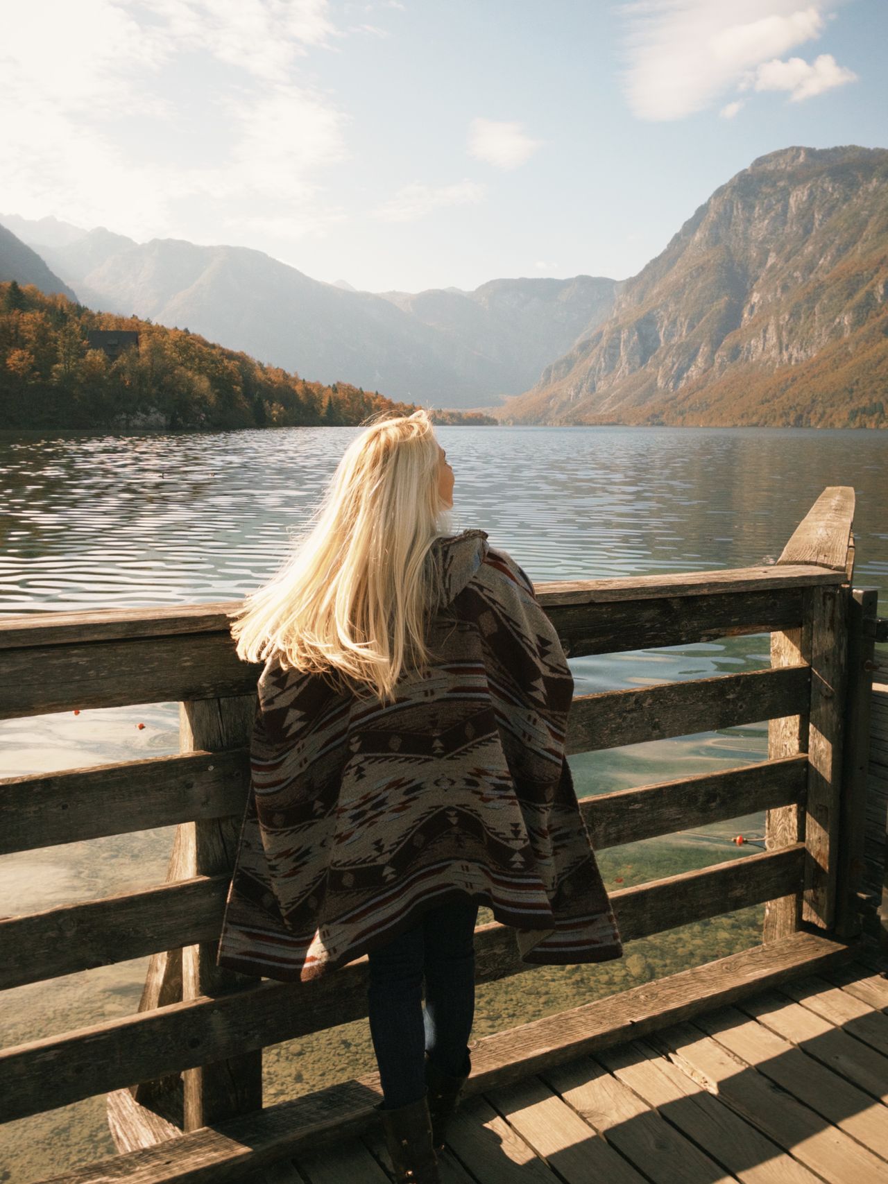 Woman wrapped in a patterned shawl standing on a wooden dock by a lake