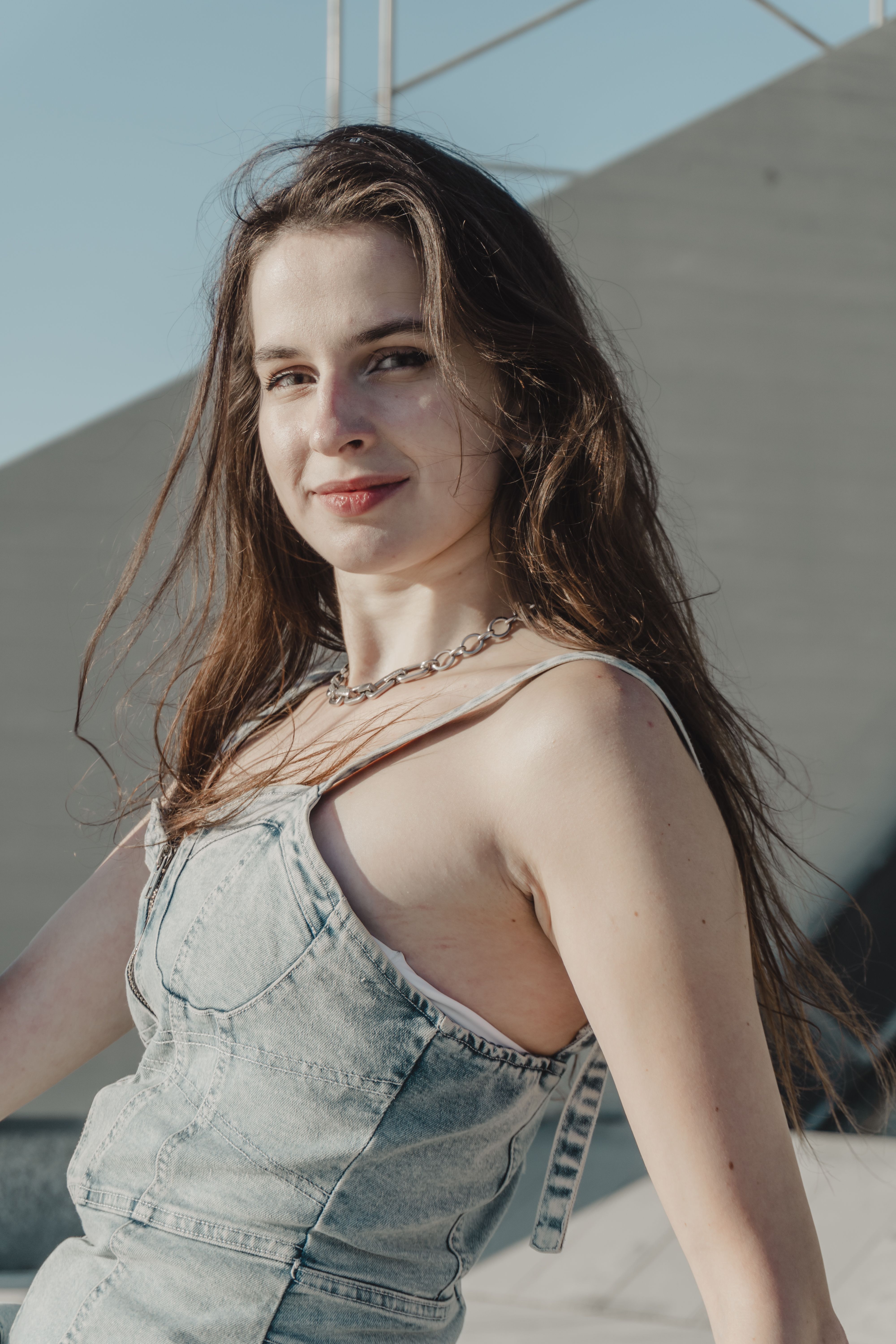 Confident young woman in a denim dress poses outdoors under bright sunlight, exuding a casual and stylish vibe.