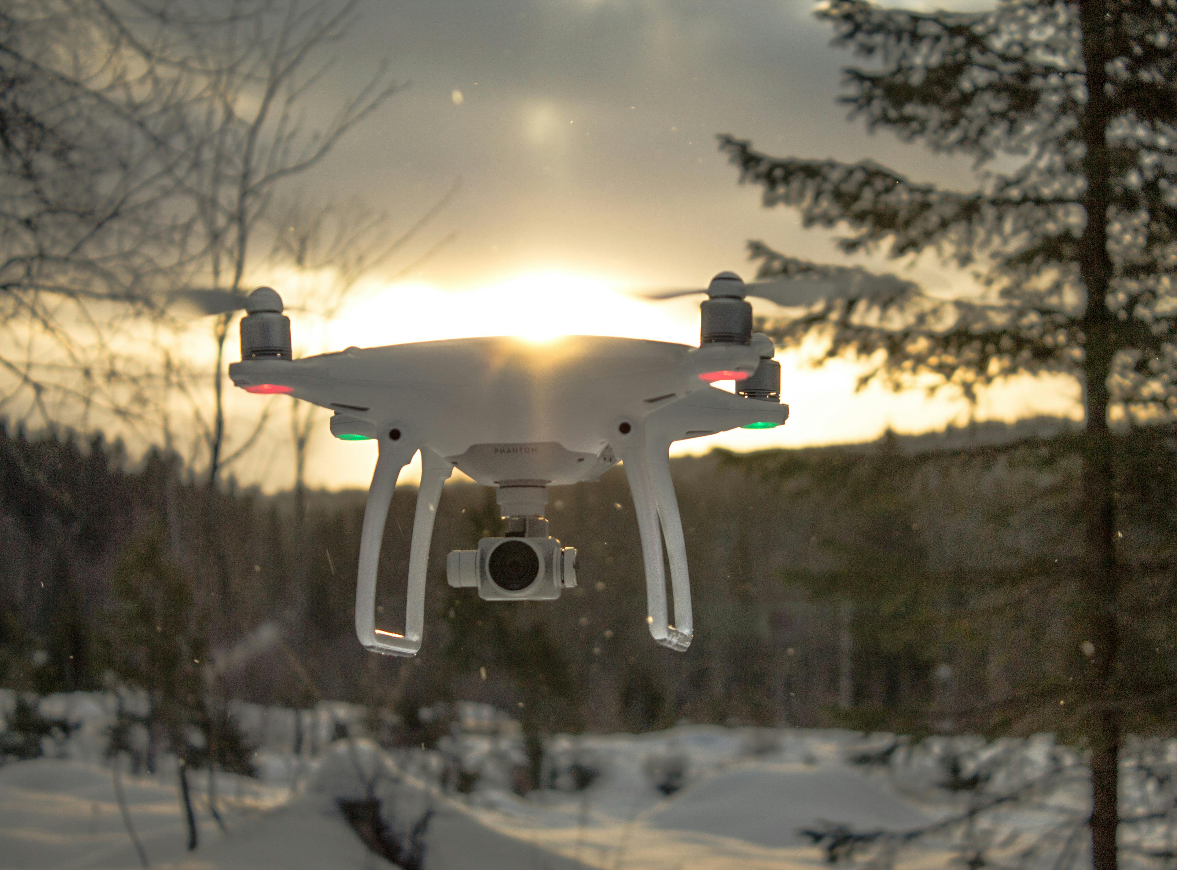 photo of white drone quadcopter flying near trees at noontime