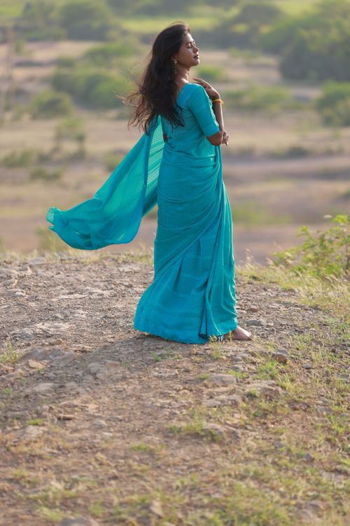Free A woman wearing a blue saree stands outdoors as the wind blows, surrounded by nature. Stock Photo