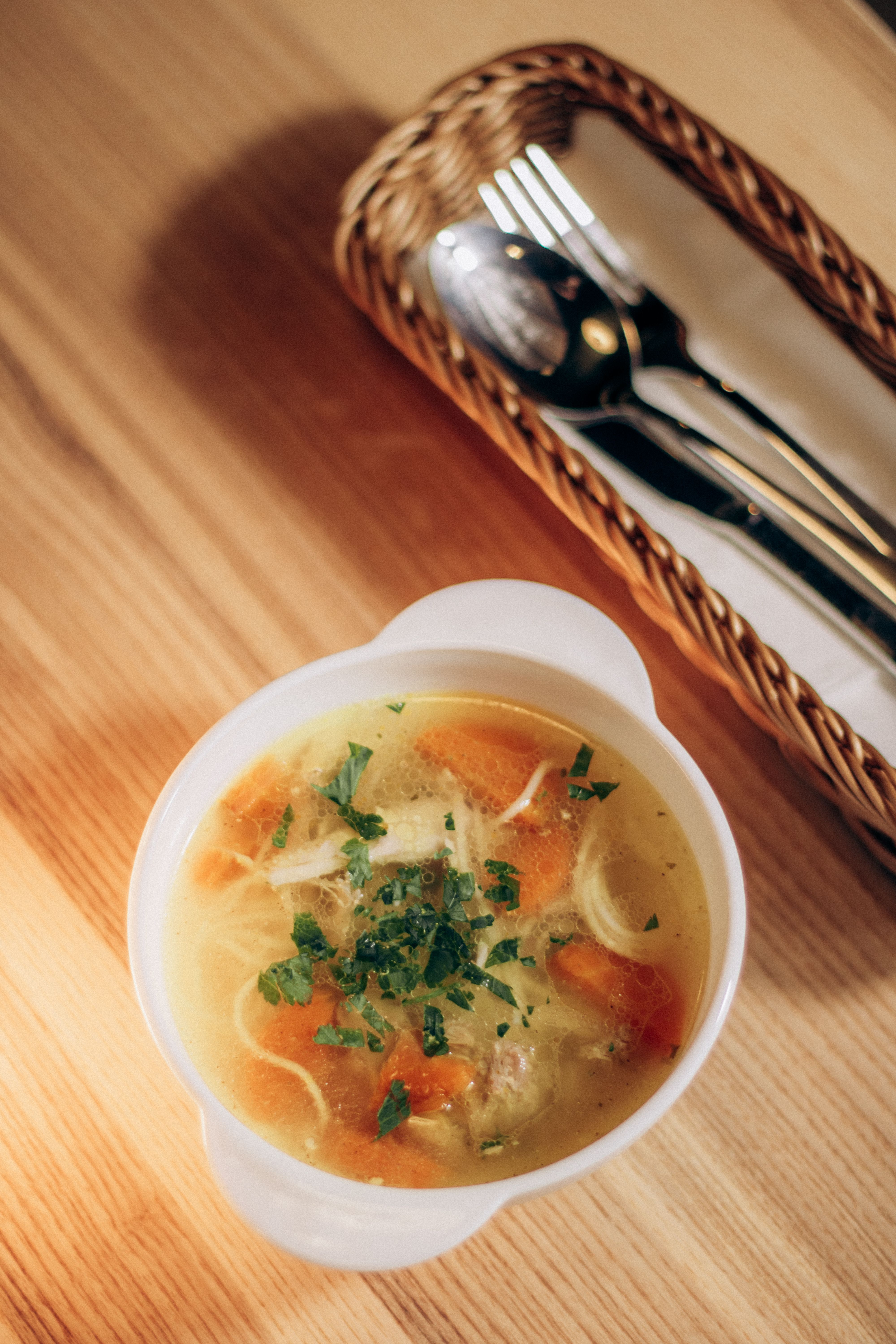 Bowl of homemade chicken soup with herbs on a wooden table beside silverware basket.