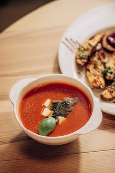 A warm tomato soup bowl garnished with basil and cheese cubes beside grilled bread.