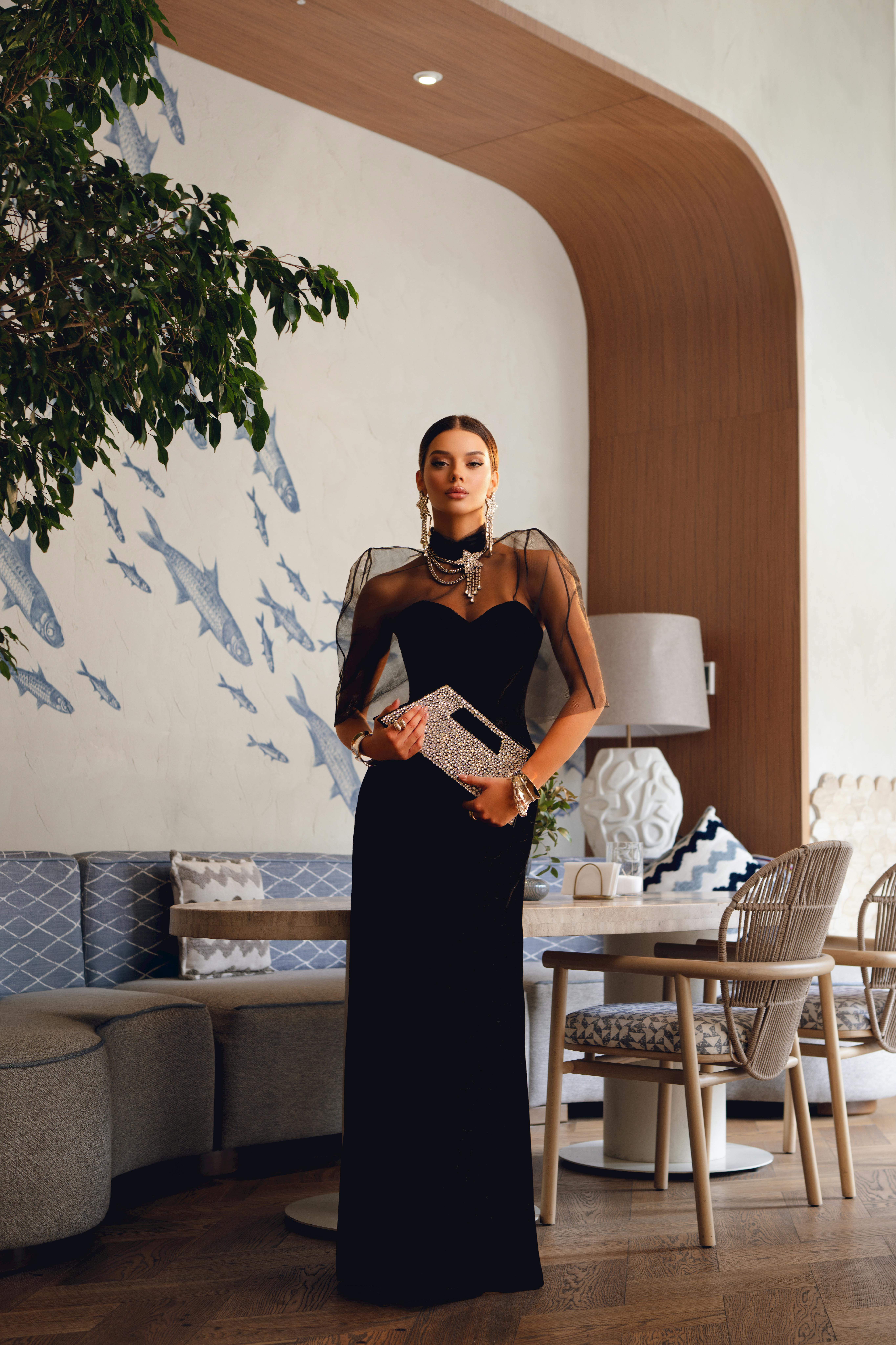 Stylish woman in a black dress poses elegantly in a modern cafe with artistic decor.