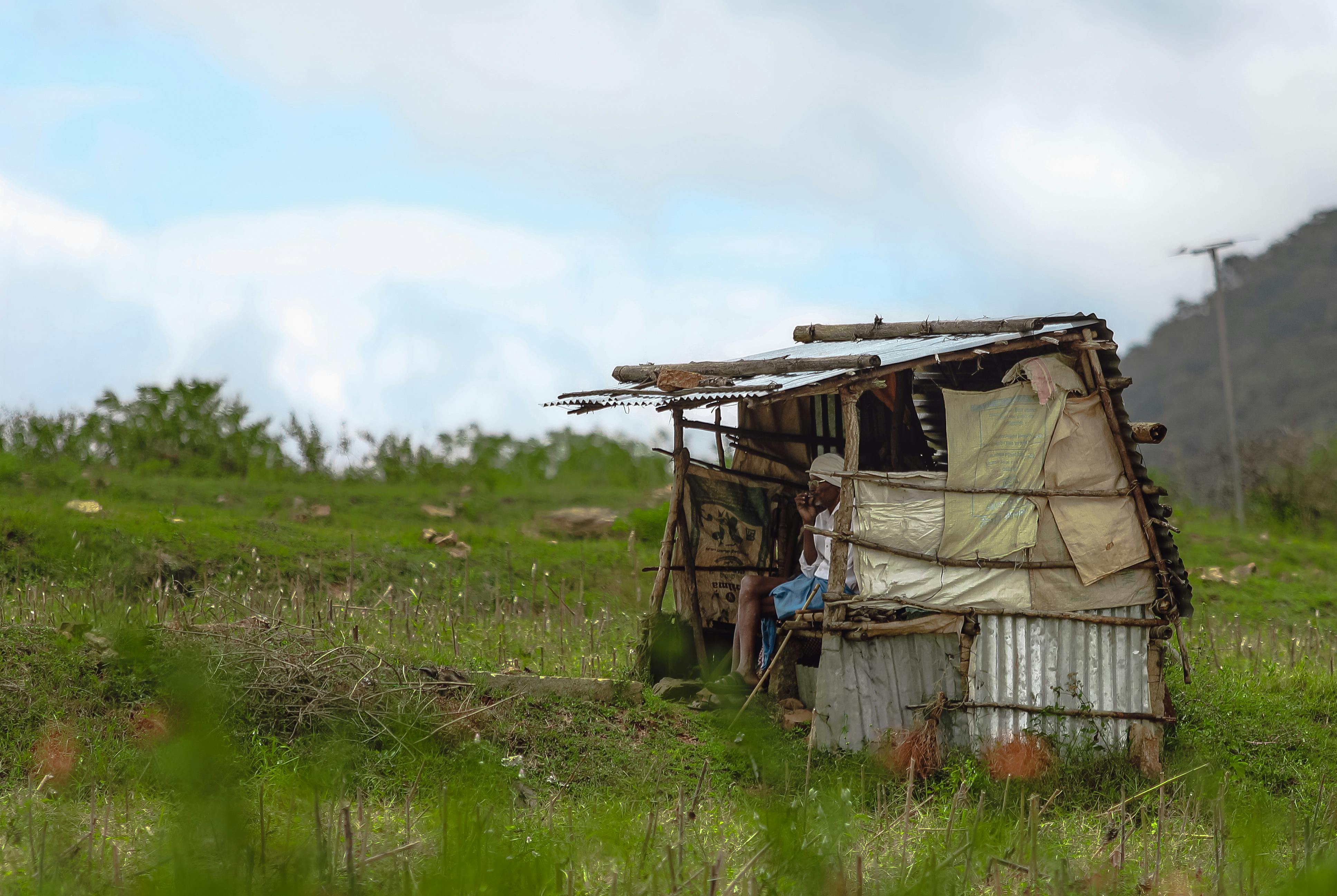 A small makeshift hut in a lush rural setting with a person inside, depicting rustic living.