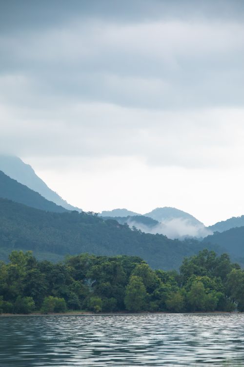 Free Peaceful view of misty mountains and calm lake reflecting greenery under cloudy sky. Stock Photo