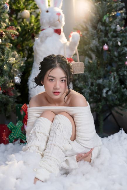 Woman in warm attire sitting among holiday decorations with a festive backdrop.