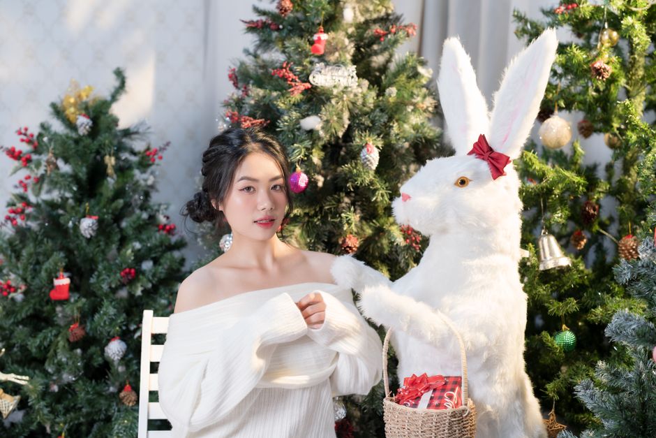 A young woman in a cozy sweater poses with a festive white rabbit amid decorated Christmas trees.