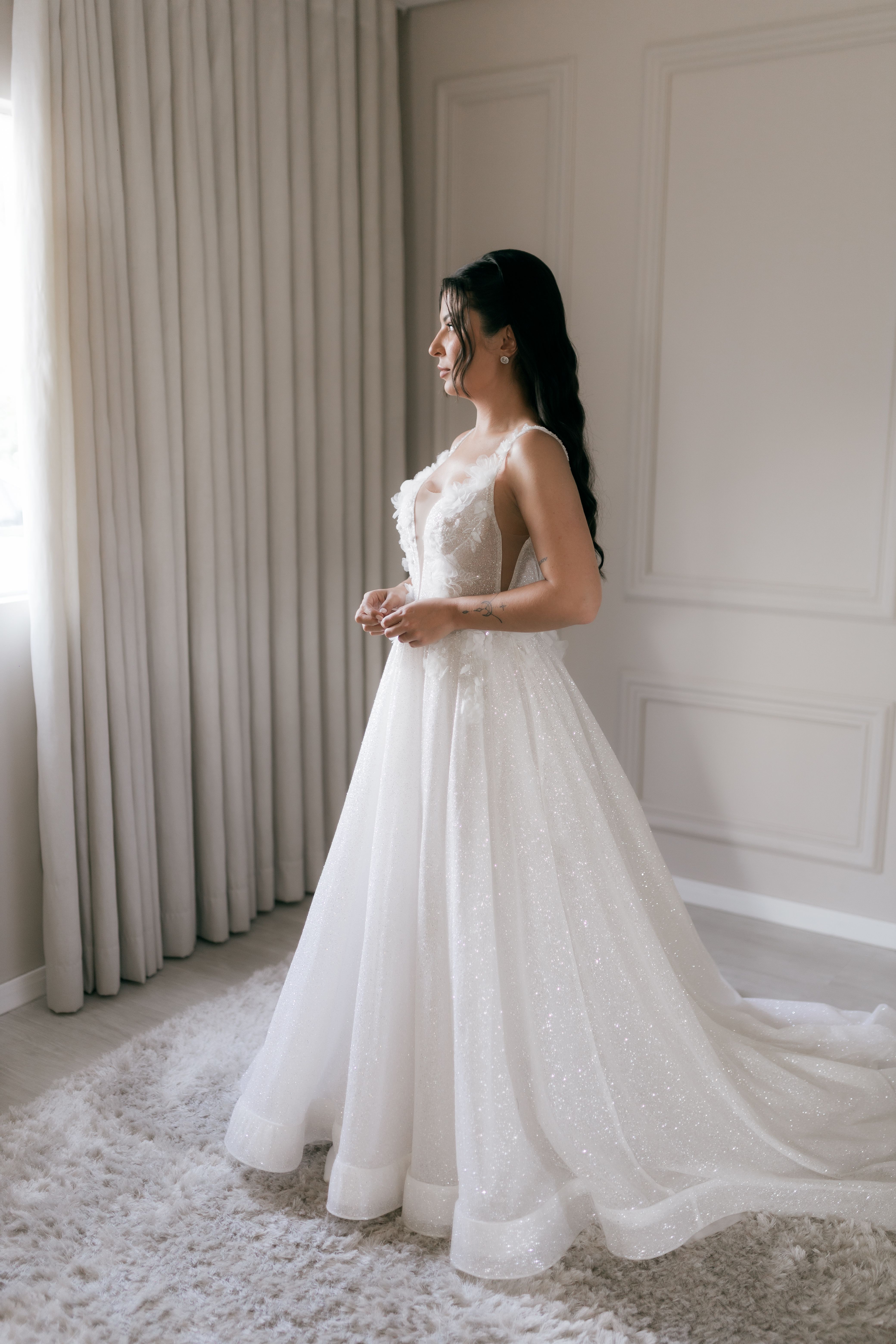 Bride in a sparkling white gown stands elegantly by a window, anticipating her big day.