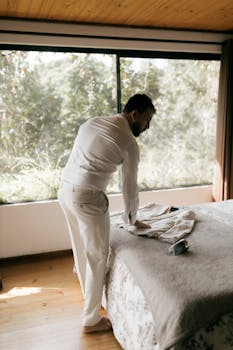 Man in white attire arranging clothes beside a large sunlit window, creating a serene indoor atmosphere.