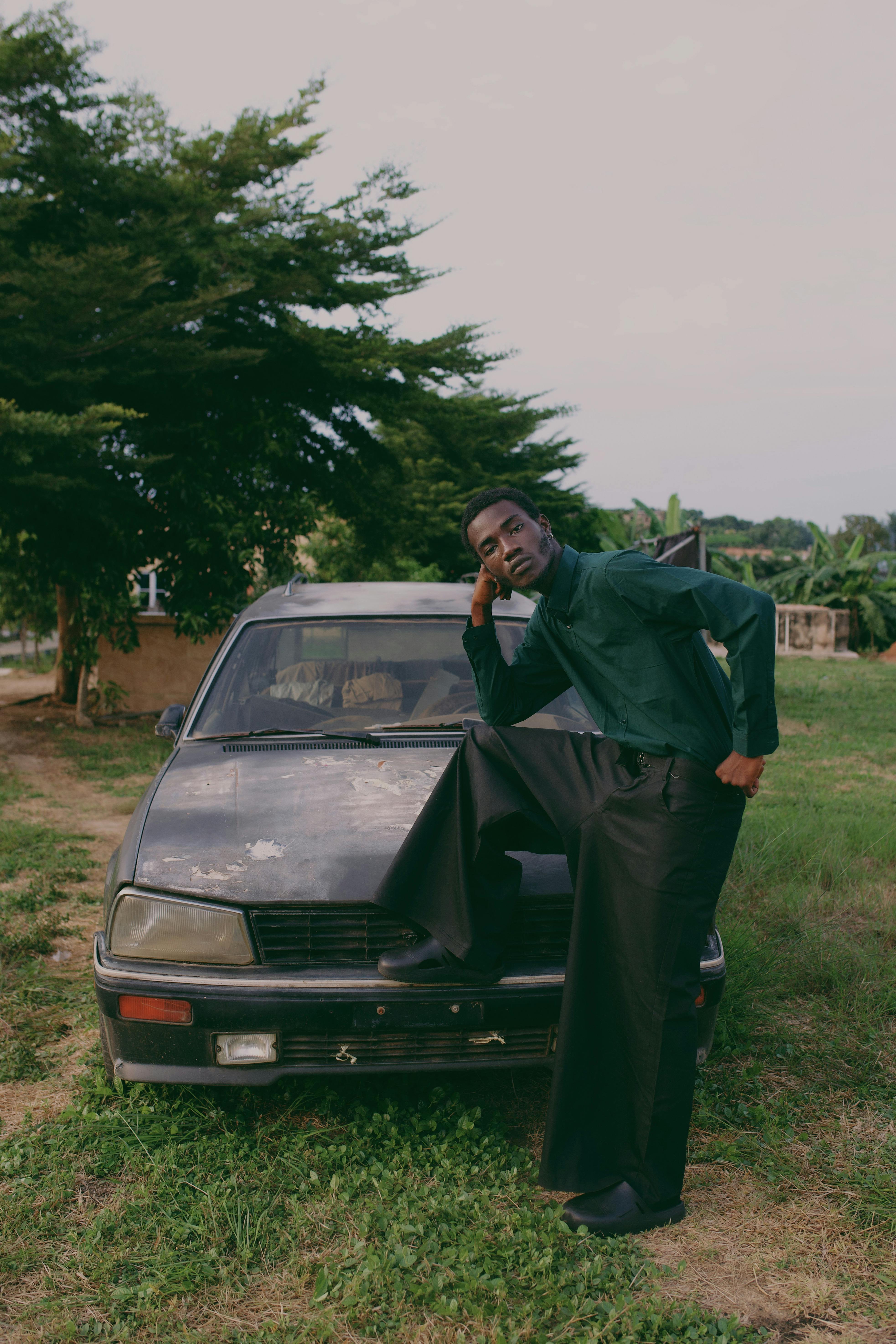 Portrait of a man casually resting on an old car in a rural Nigerian setting.