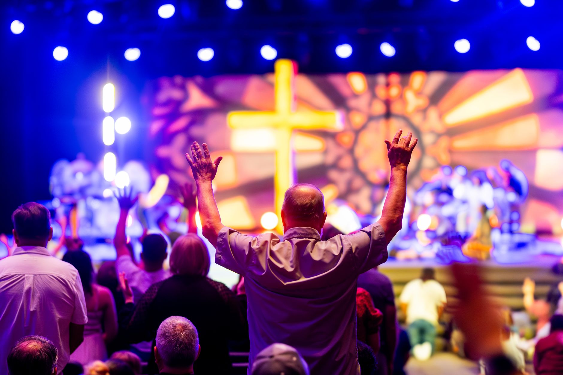 A lively worship service with people praising, colorful lights, and a cross stage backdrop.