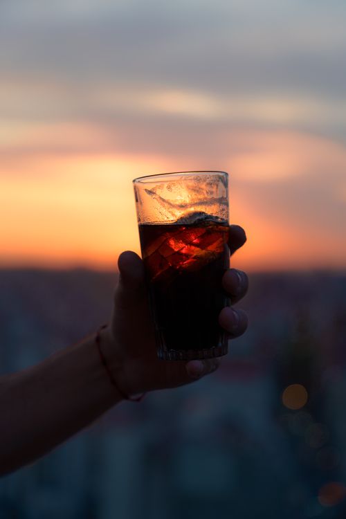 Free A hand holds a refreshing cold drink in a glass against a beautiful sunset backdrop. Stock Photo
