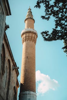 A towering historic minaret reaches up into the clear blue sky, framed by lush foliage.