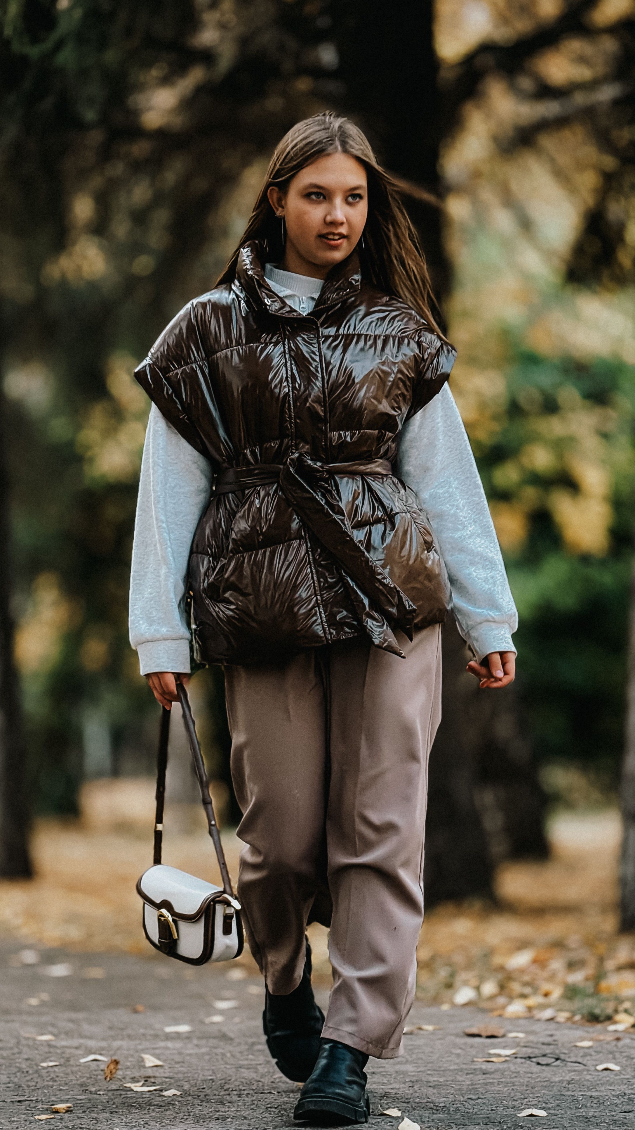 Young woman in stylish fall outfit walking in park, carrying handbag.